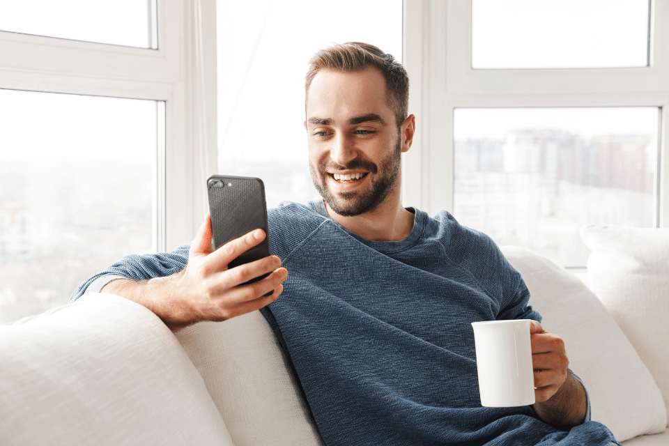 Attractive young man relaxing on a couch at home while drinking coffee, and using his mobile phone to talk to his Filipina girlfriend.