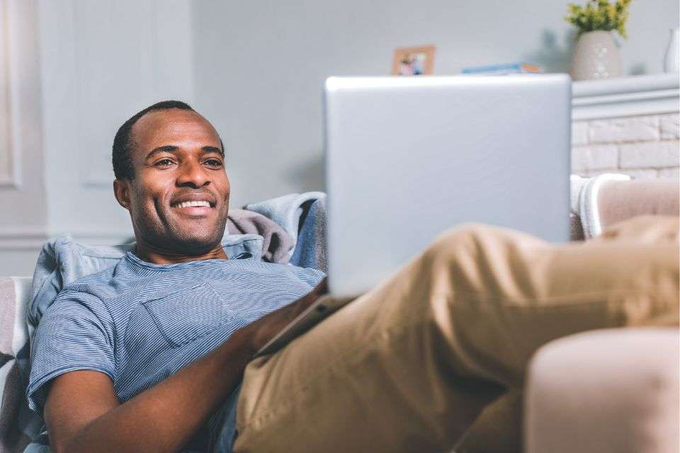 An African American Man Using His Laptop to Go Dating Online while Lying on the Sofa